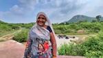 Smiling woman in colorful saree near a pond with lush greenery and a hill in the background.