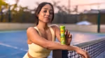A woman in a yellow sports outfit stands on a tennis court at sunset, leaning on a net post. She's holding a green Lipton Energy Boost bottle, smiling slightly.